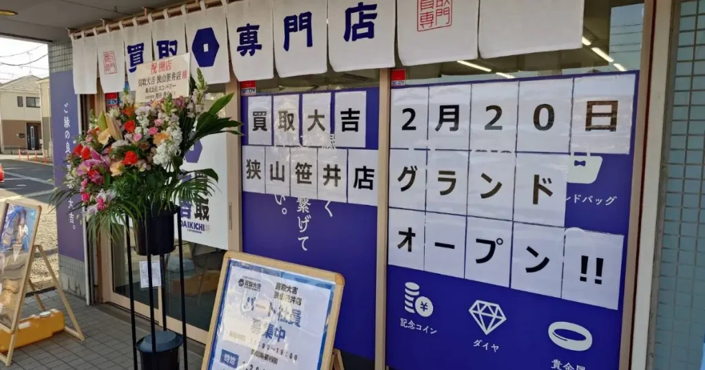 2月20日グランドオープンと書かれた大きな貼り紙がある買取大吉狭山笹井店の店舗外観と入り口の祝い花