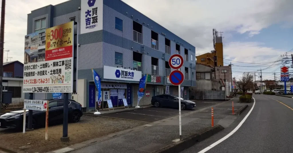 買取大吉の青い看板を掲げた3階建ての建物と手前に設置されたミライズ株式会社の不動産広告看板がある道路沿いの風景