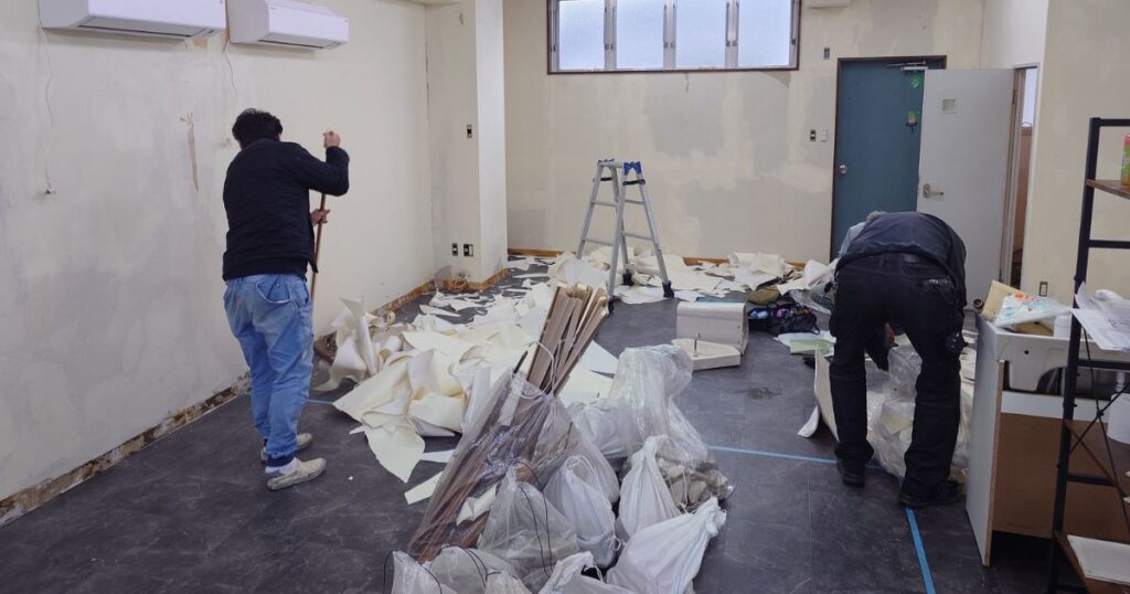 剥がした壁紙やゴミが散乱する部屋で改装作業をする2人の作業員